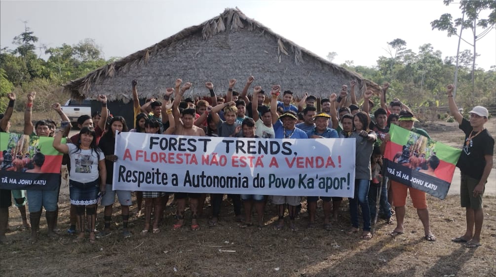 Con le braccia tese e i pugni stretti, più di 30 persone di Ka'apor protestano davanti a una casa con pilastri di legno e tetto di paglia. Alcuni di loro reggono uno striscione che recita: "Forest Trends, la foresta non è in vendita".