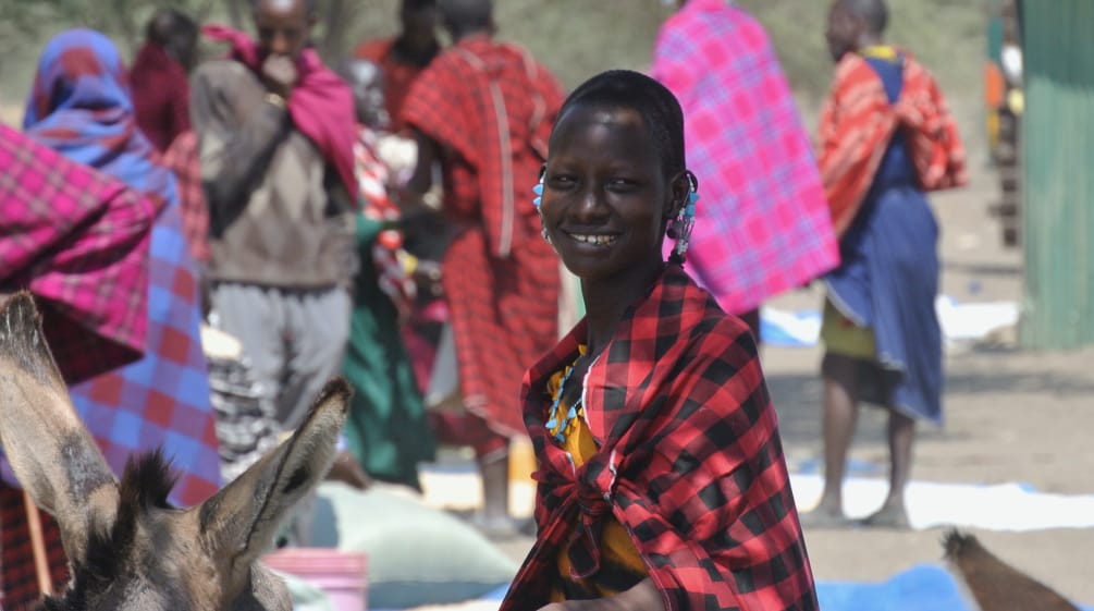 Donne e uomini Maasai in abiti colorati in un mercato, una donna guarda nell'obiettivo