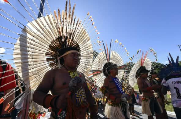 Tre uomini indigeni con grandi copricapi rotondi di piume in testa, con in mano le loro maracas