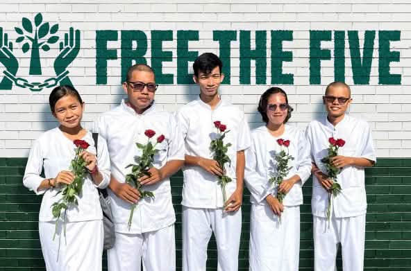Cinque persone vestite di bianco, con in mano rose rosse, si trovano davanti a un muro su cui si legge: «Liberate i Cinque».