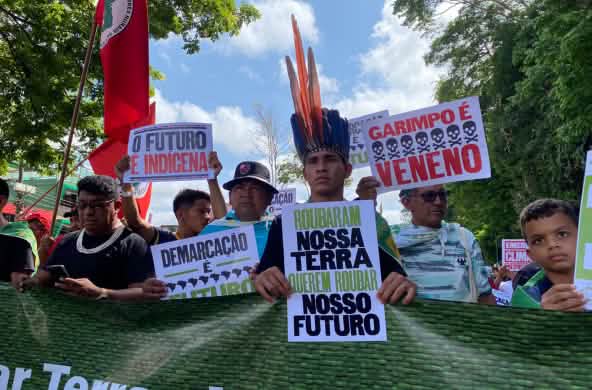Indigeni che marciano in fila reggendo cartelli con slogan come: “La nostra terra è il nostro futuro”