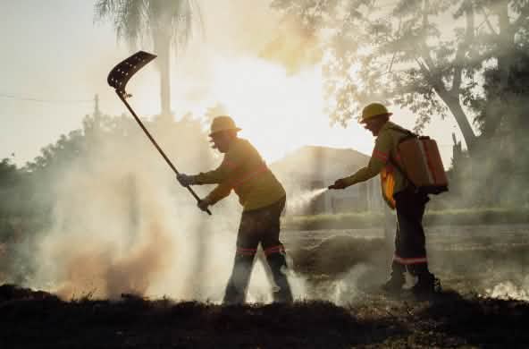 Tra il fumo, un uomo combatte il fuoco con un battifuoco, mentre un altro uomo porta uno zaino idrico e una manichetta manuale