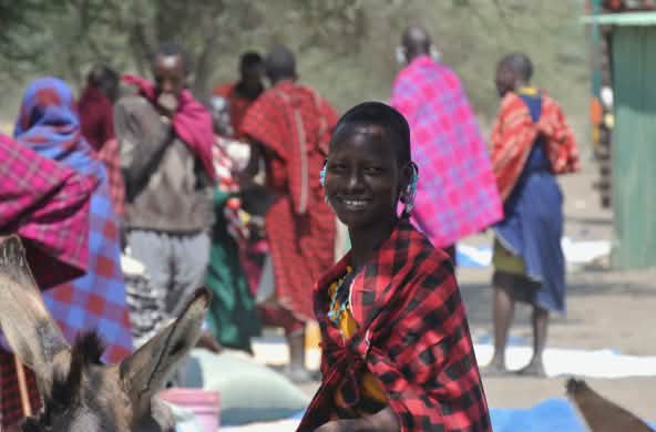 Donne e uomini Maasai in abiti colorati in un mercato, una donna guarda nell'obiettivo
