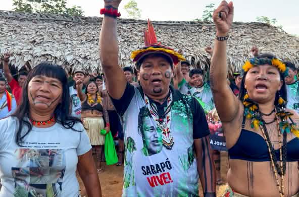Tre indigeni Ka'apor - due donne e un uomo, con i pugni alzati davanti a uno spazio coperto da foglie di palma.