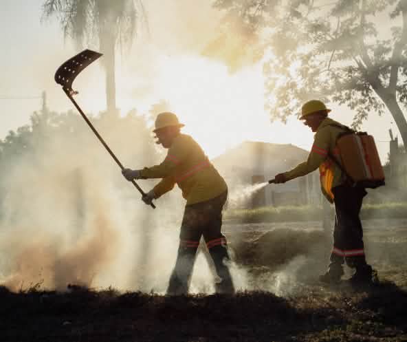 Tra il fumo, un uomo combatte il fuoco con un battifuoco, mentre un altro uomo porta uno zaino idrico e una manichetta manuale