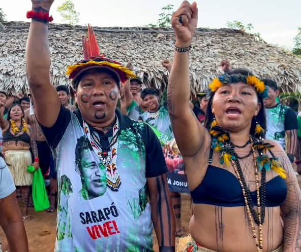 Tre indigeni Ka'apor - due donne e un uomo, con i pugni alzati davanti a uno spazio coperto da foglie di palma.