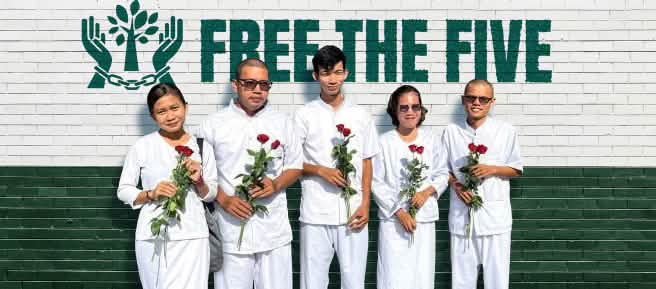 Cinque persone vestite di bianco, con in mano rose rosse, si trovano davanti a un muro su cui si legge: «Liberate i Cinque».