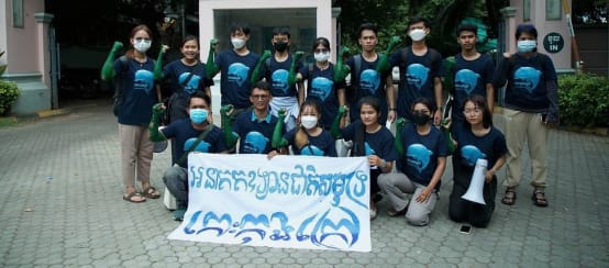 15 giovani sono in piedi e in ginocchio davanti all'edificio del Ministero del Turismo e tengono uno striscione verso la macchina fotografica. Lo striscione recita: "L'isola di Koh Kong deve diventare un parco nazionale marino".
