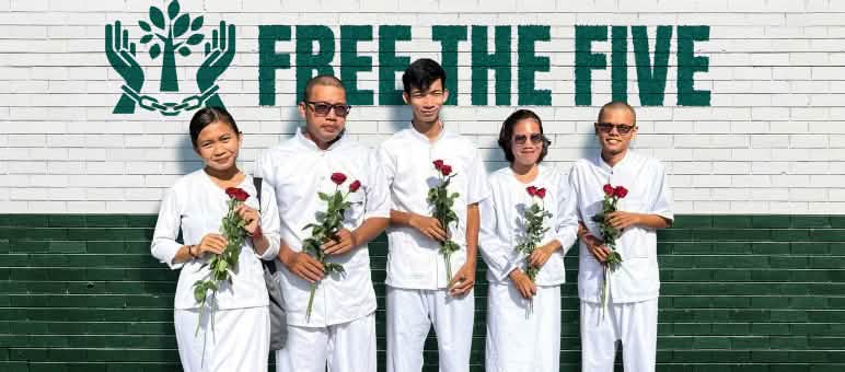 Cinque persone vestite di bianco, con in mano rose rosse, si trovano davanti a un muro su cui si legge: «Liberate i Cinque».