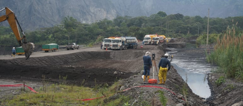 Gli ingegneri ispezionano il terreno tra una fossa di terra piena di petrolio greggio sulla destra. Sulla sinistra un escavatore, che crea un'altra fossa di terra. Sullo sfondo, si vedono delle autocisterne davanti alla montagna.