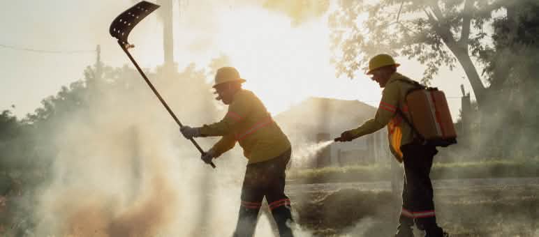 Tra il fumo, un uomo combatte il fuoco con un battifuoco, mentre un altro uomo porta uno zaino idrico e una manichetta manuale