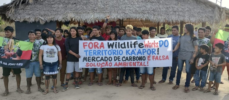 Più di 30 indigeni Ka'apor si trovano davanti a una casa tradizionale con pilastri di legno e tetto di paglia. Le persone reggono uno striscione di protesta che recita: "Wildlife Works fuori dal territorio Ka'apor! Il mercato del carbonio è un falso sole