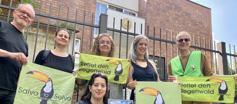 6 persone sono in piedi davanti alla grata dell'edificio dell'ambasciata, con in mano striscioni con le scritte Rettet den Regenwald e Salva la Selva con il tucano simbolo dell'associazione.