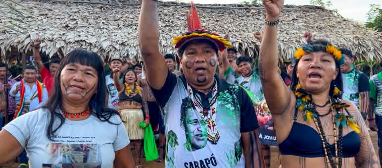 Tre indigeni Ka'apor - due donne e un uomo, con i pugni alzati davanti a uno spazio coperto da foglie di palma.