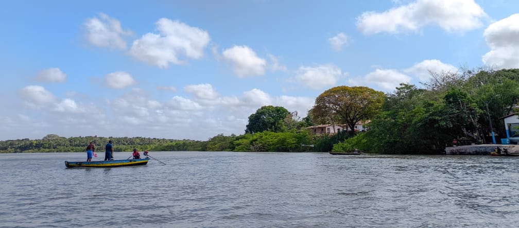 Sotto un cielo azzurro, tre pescatori su una piccola imbarcazione navigano al largo. A destra si vedono alcune piccole case sulla costa, tra la vegetazione.