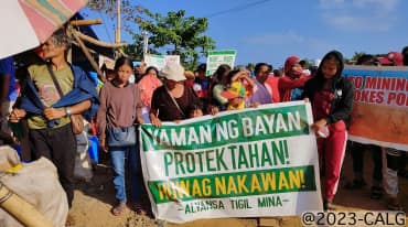 Un gruppo di persone regge un cartello con la scritta (tradotta): Proteggere la ricchezza del popolo! No ai saccheggi! Alleanza contro l'estrazione mineraria.
