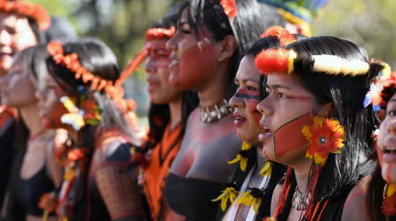 Protesta indigena all'Accampamento della Terra Libera a Brasilia 2024 Otto donne indigene, una accanto all'altra, con copricapi di piume, orecchini di piume gialle e rosse e pittura facciale rossa e nera.