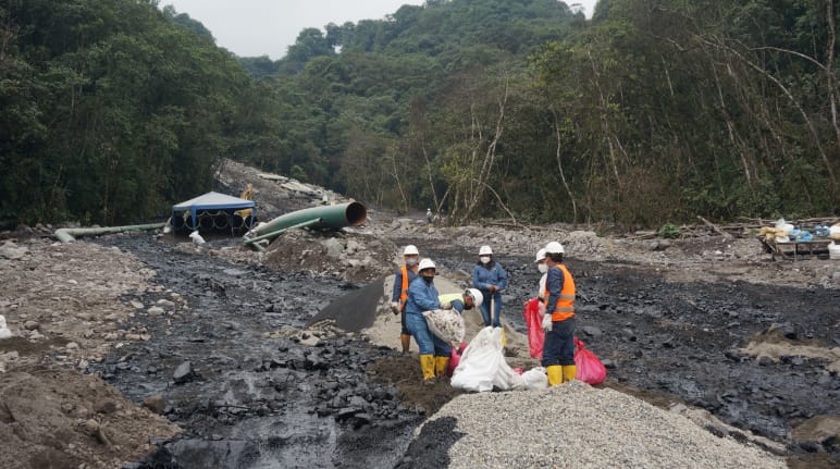 Un gruppo di lavoratori petroliferi lavora tra due fiumi neri di petrolio che scendono da una montagna coperta di rocce nella foresta. Una sezione di tubo dell'oleodotto si vede sullo sfondo.