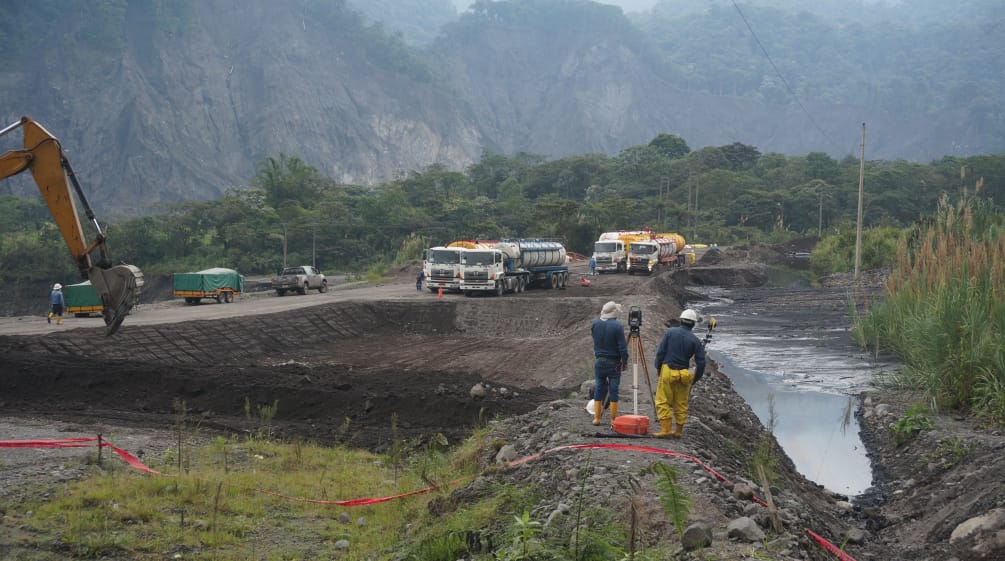 Gli ingegneri ispezionano il terreno tra una fossa di terra piena di petrolio greggio sulla destra. Sulla sinistra un escavatore, che crea un'altra fossa di terra. Sullo sfondo, si vedono delle autocisterne davanti alla montagna.