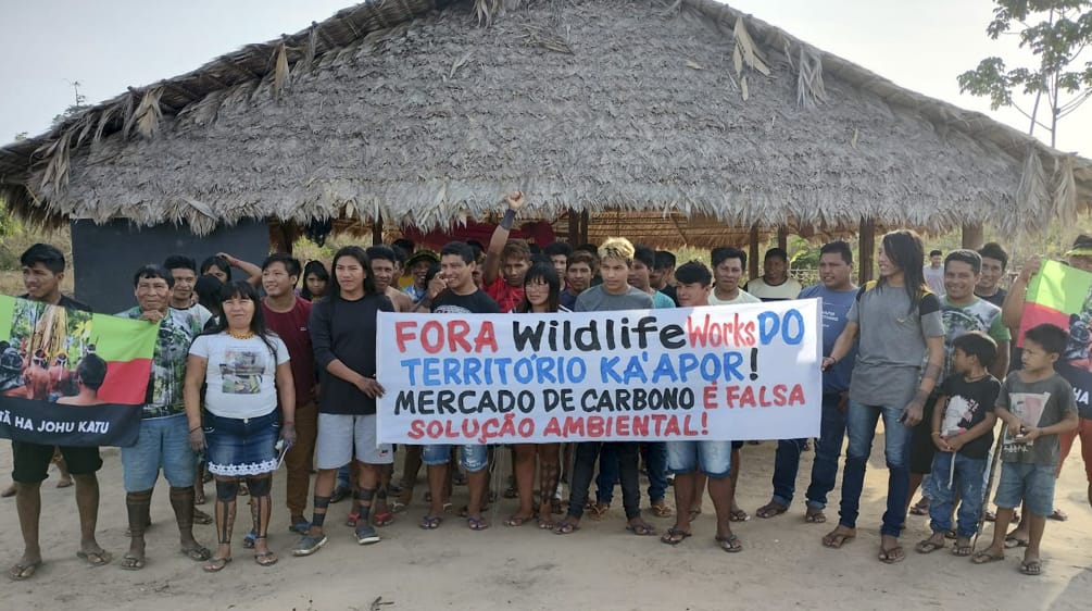 Più di 30 indigeni Ka'apor si trovano davanti a una casa tradizionale con pilastri di legno e tetto di paglia. Le persone reggono uno striscione di protesta che recita: "Wildlife Works fuori dal territorio Ka'apor! Il mercato del carbonio è un falso sole