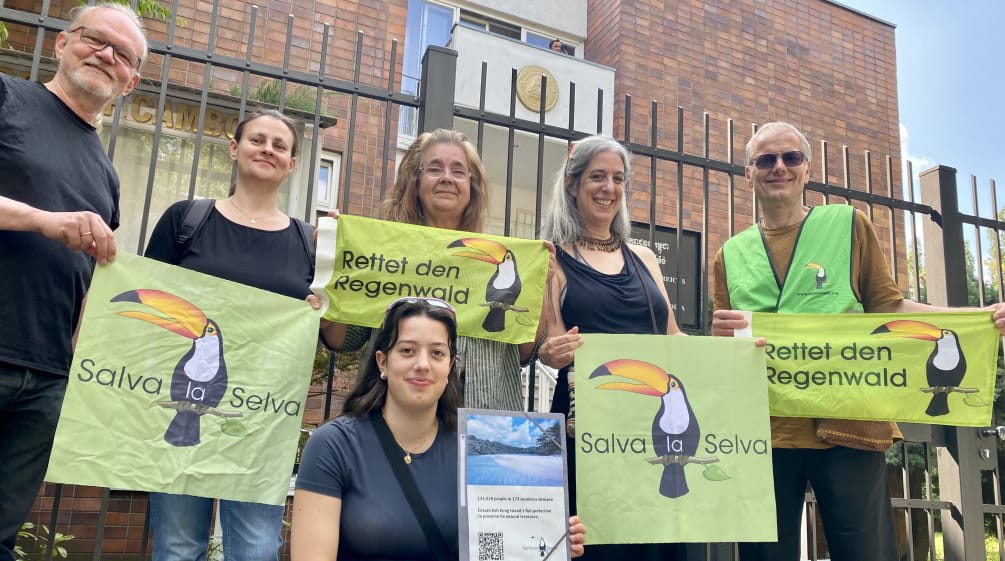 6 persone sono in piedi davanti alla grata dell'edificio dell'ambasciata, con in mano striscioni con le scritte Rettet den Regenwald e Salva la Selva con il tucano simbolo dell'associazione.