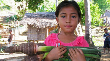 Una bambina di Palwan ha tra le braccia il Taro, un vegetale locale, e sullo sfondo si intravede il villaggio